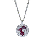 A silver pendant necklace featuring a textured circular disc with three faceted deep red gemstones, suspended from a silver chain on a plain white background.