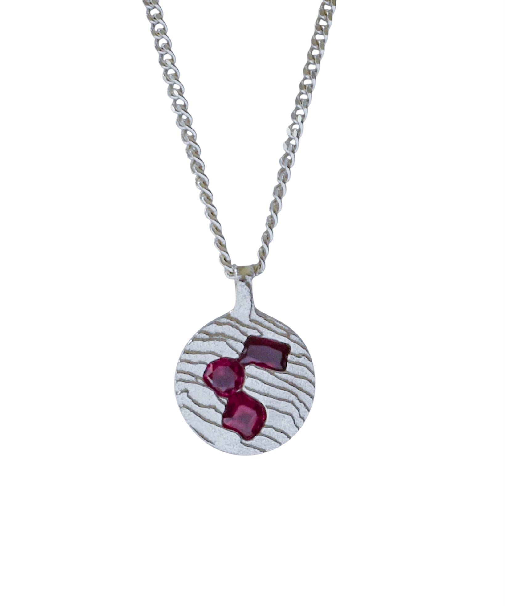 A silver pendant necklace featuring a textured circular disc with three faceted deep red gemstones, suspended from a silver chain on a plain white background.