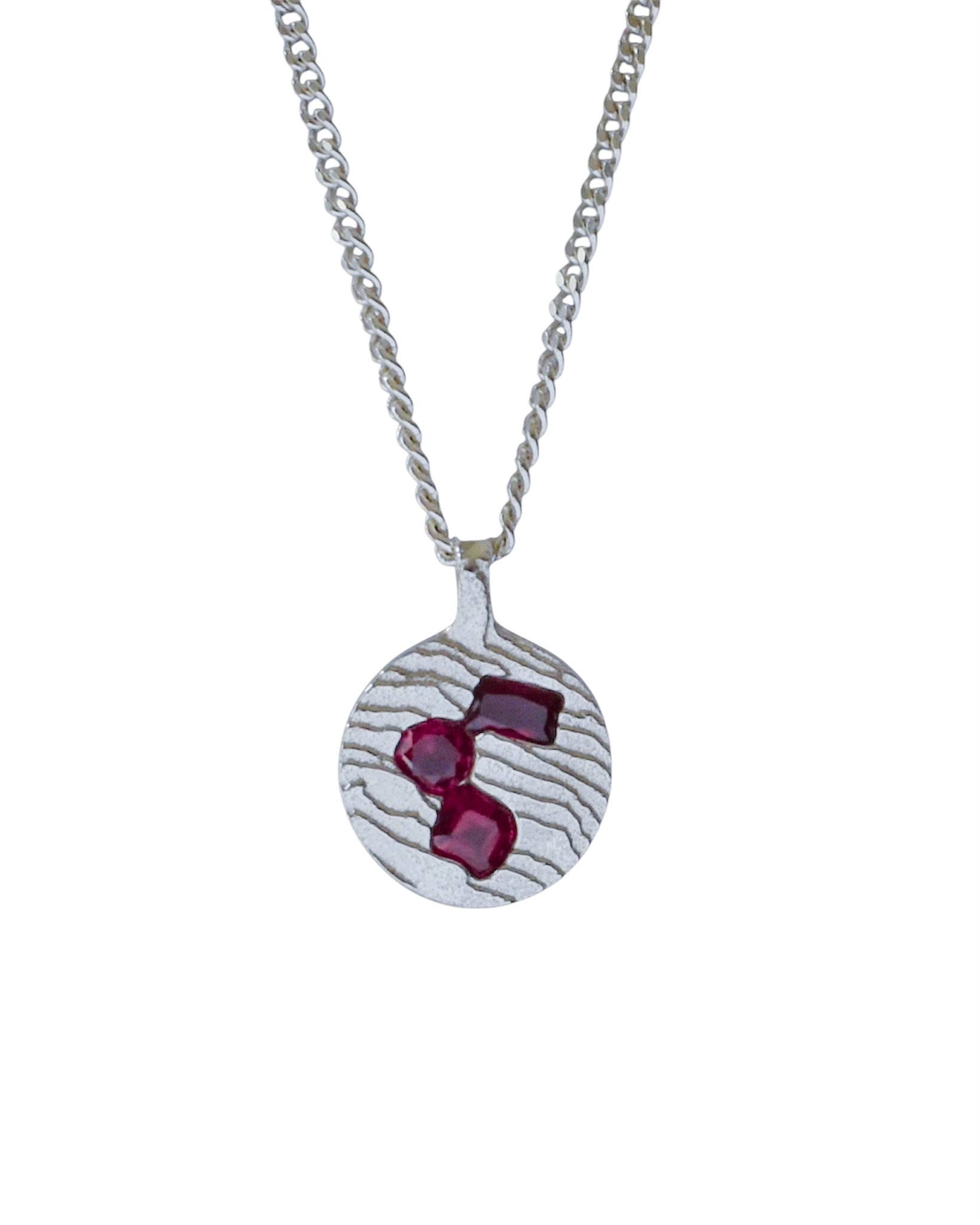 A silver pendant necklace featuring a textured circular disc with three faceted deep red gemstones, suspended from a silver chain on a plain white background.