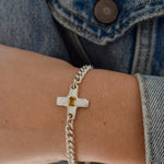 A close-up of a person's wrist wearing a silver chain bracelet with a textured cross charm featuring a small yellow gemstone, styled with a blue denim jacket.