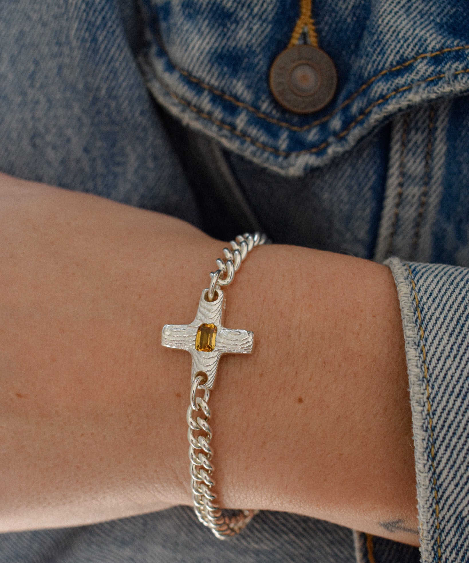A close-up of a person's wrist wearing a silver chain bracelet with a textured cross charm featuring a small yellow gemstone, styled with a blue denim jacket.
