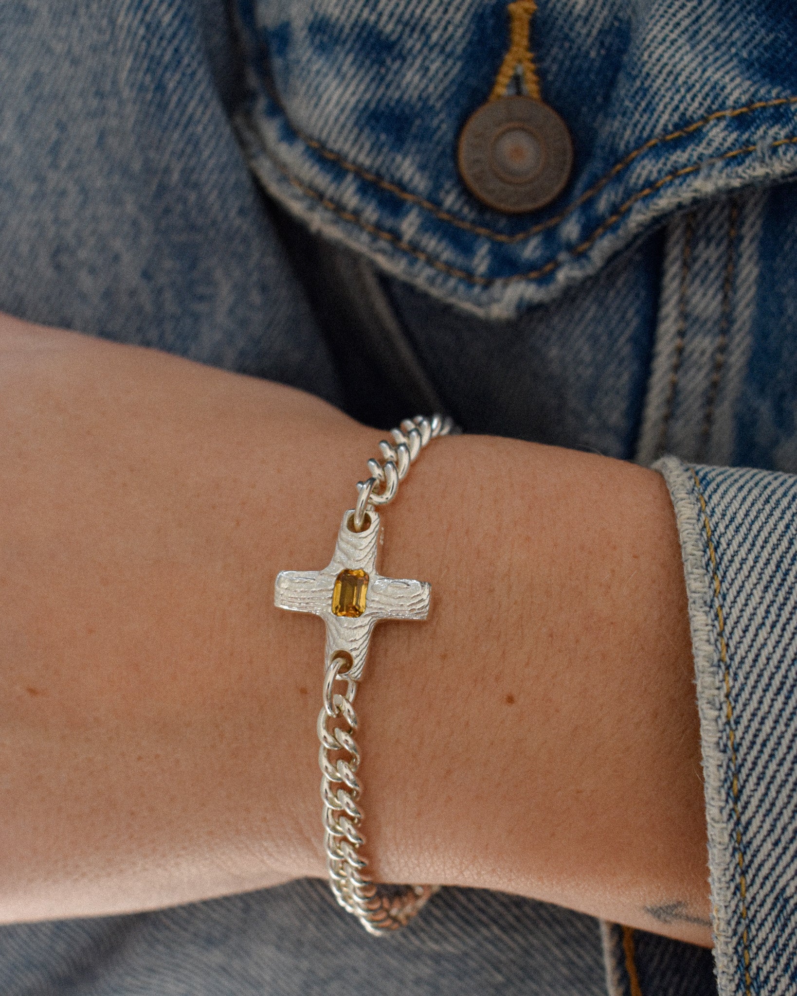 A close-up of a person's wrist wearing a silver chain bracelet with a textured cross charm featuring a small yellow gemstone, styled with a blue denim jacket.