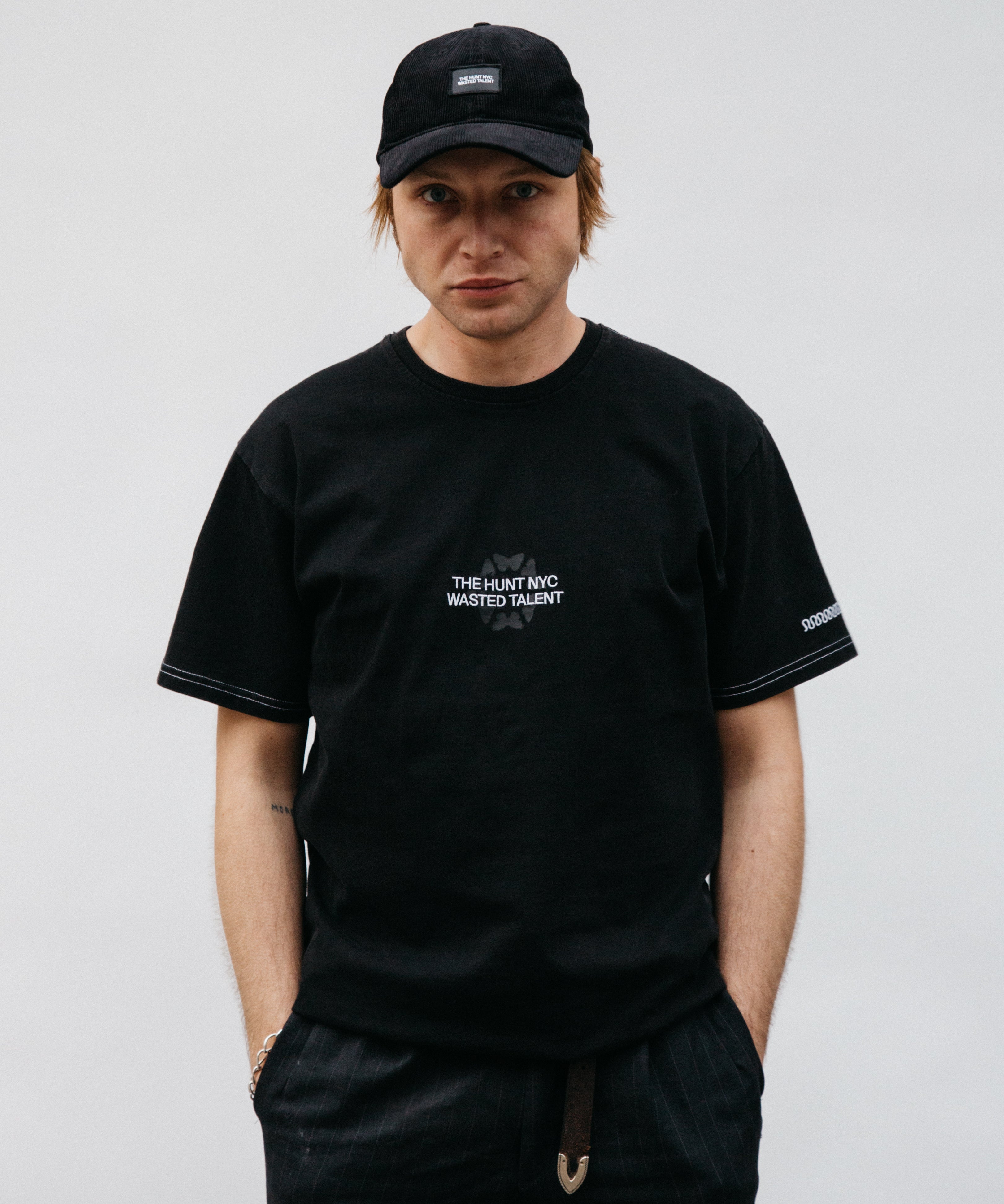 Streetwear model wearing black Wasted Talent x THE HUNT NYC t-shirt with embroidered butterfly graphic and black cap — front-facing product lifestyle image.