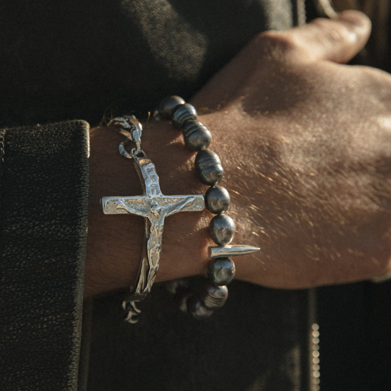 Close-up of a wrist wearing a silver cross bracelet with dark beads on a dark background