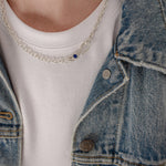 A close-up of a person wearing a delicate silver chain necklace with a small dark blue oval stone pendant, styled over a white t-shirt and a blue denim jacket.