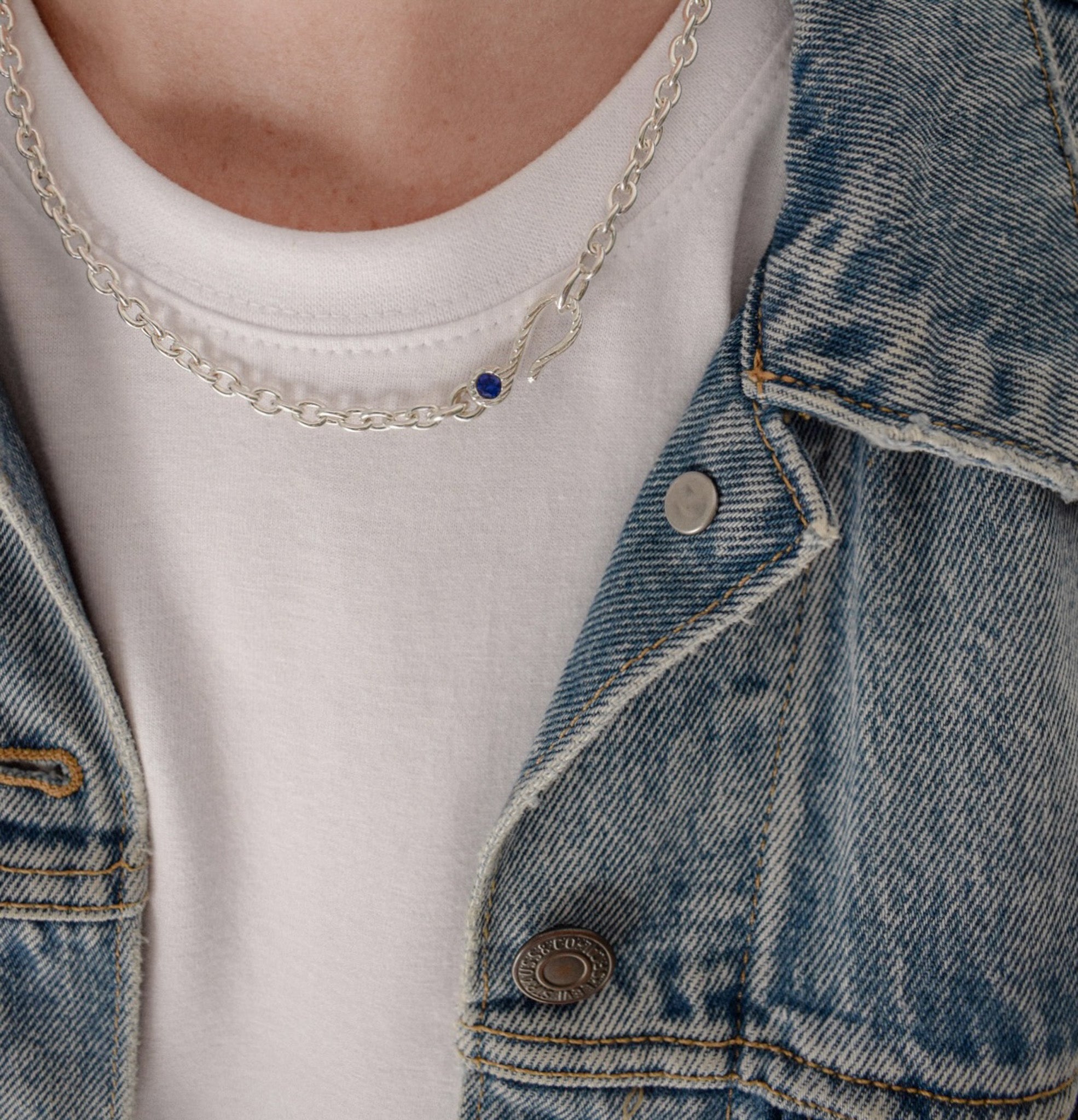 A close-up of a person wearing a delicate silver chain necklace with a small dark blue oval stone pendant, styled over a white t-shirt and a blue denim jacket.