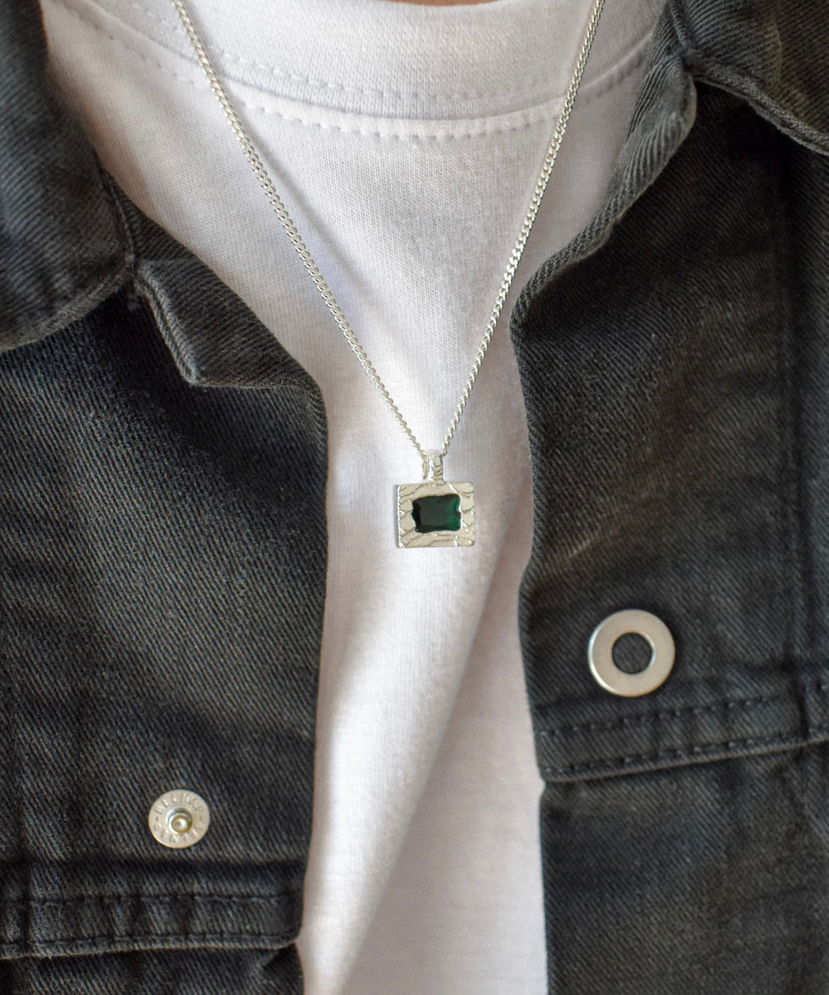A close-up of a person wearing a silver chain necklace with a square pendant featuring a dark green center stone, styled over a white t-shirt and a dark denim jacket.