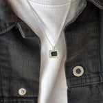A close-up of a person wearing a silver chain necklace with a square pendant featuring a dark green center stone, styled over a white t-shirt and a dark denim jacket.