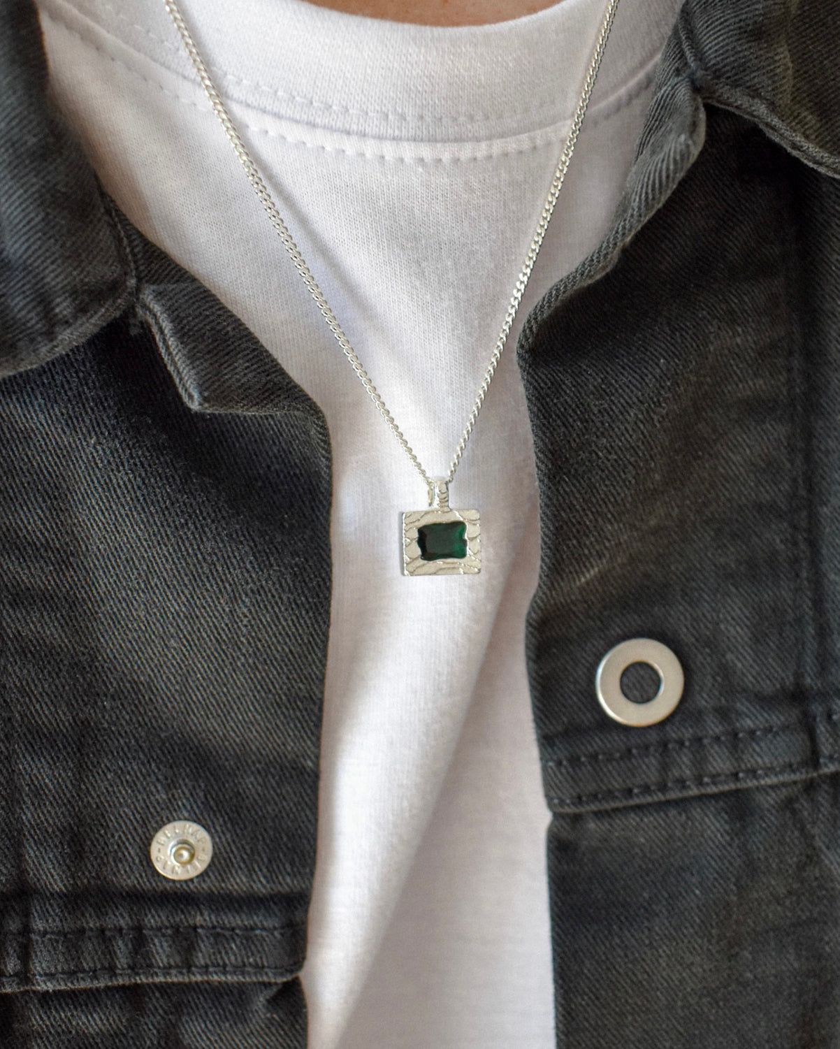A close-up of a person wearing a silver chain necklace with a square pendant featuring a dark green center stone, styled over a white t-shirt and a dark denim jacket.