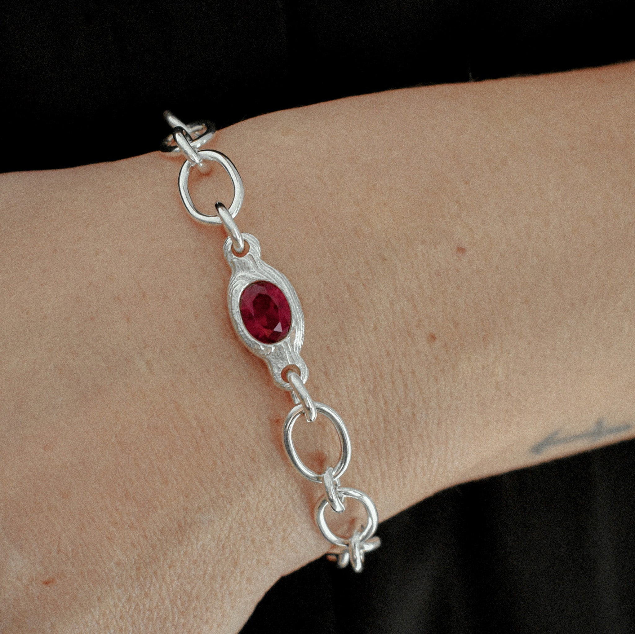 A sterling silver chain bracelet with a central oval red gemstone, worn on a person's wrist against a black background.