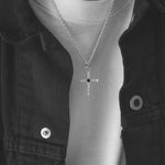 A silver cross pendant necklace with a small black center stone, worn on a model's neck over a white t-shirt and dark denim jacket.