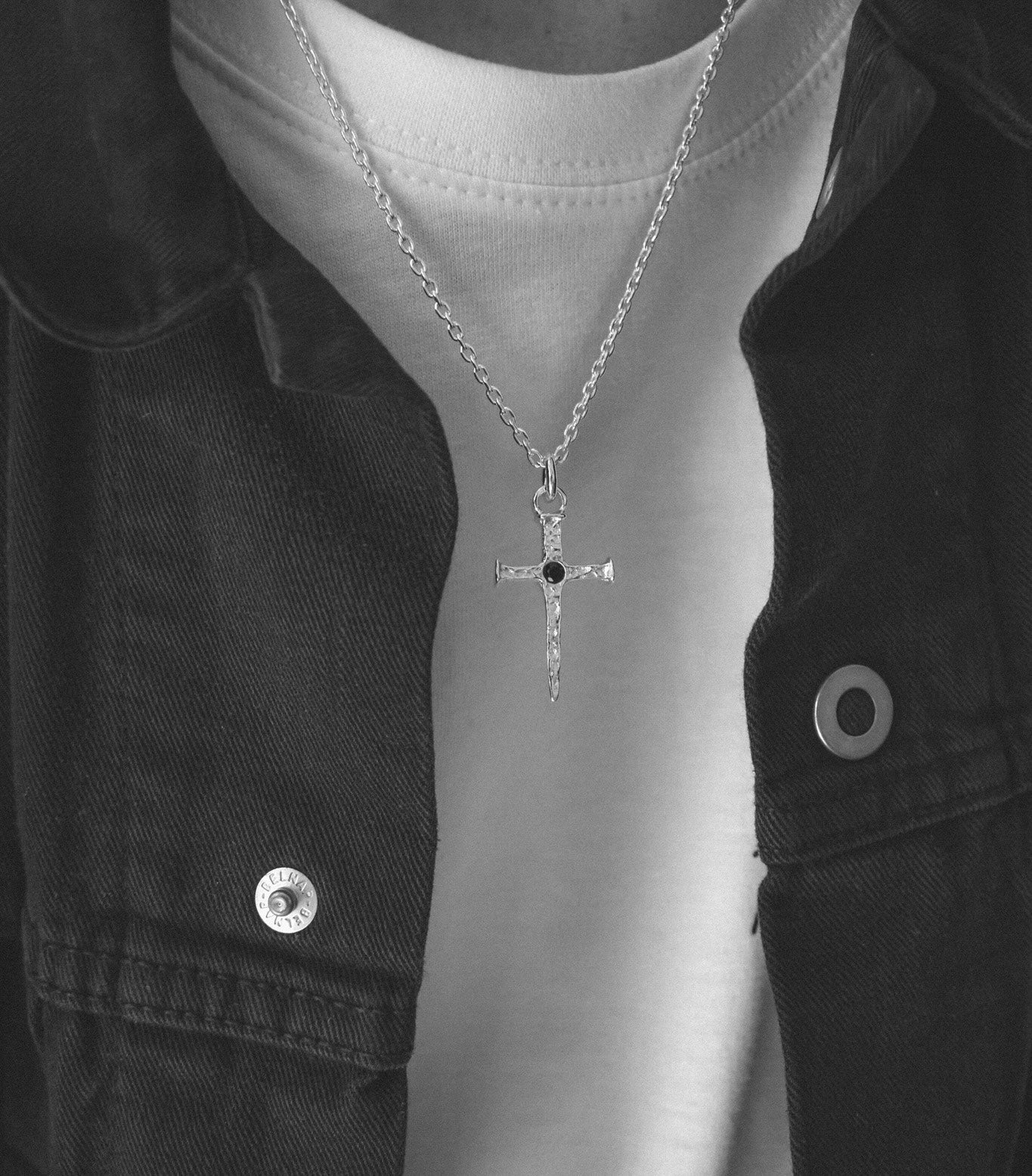 A silver cross pendant necklace with a small black center stone, worn on a model's neck over a white t-shirt and dark denim jacket.