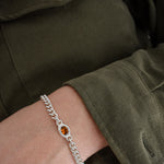 A close-up of a person's wrist wearing a silver chain bracelet with a central oval amber gemstone, set against an olive green military-style jacket.