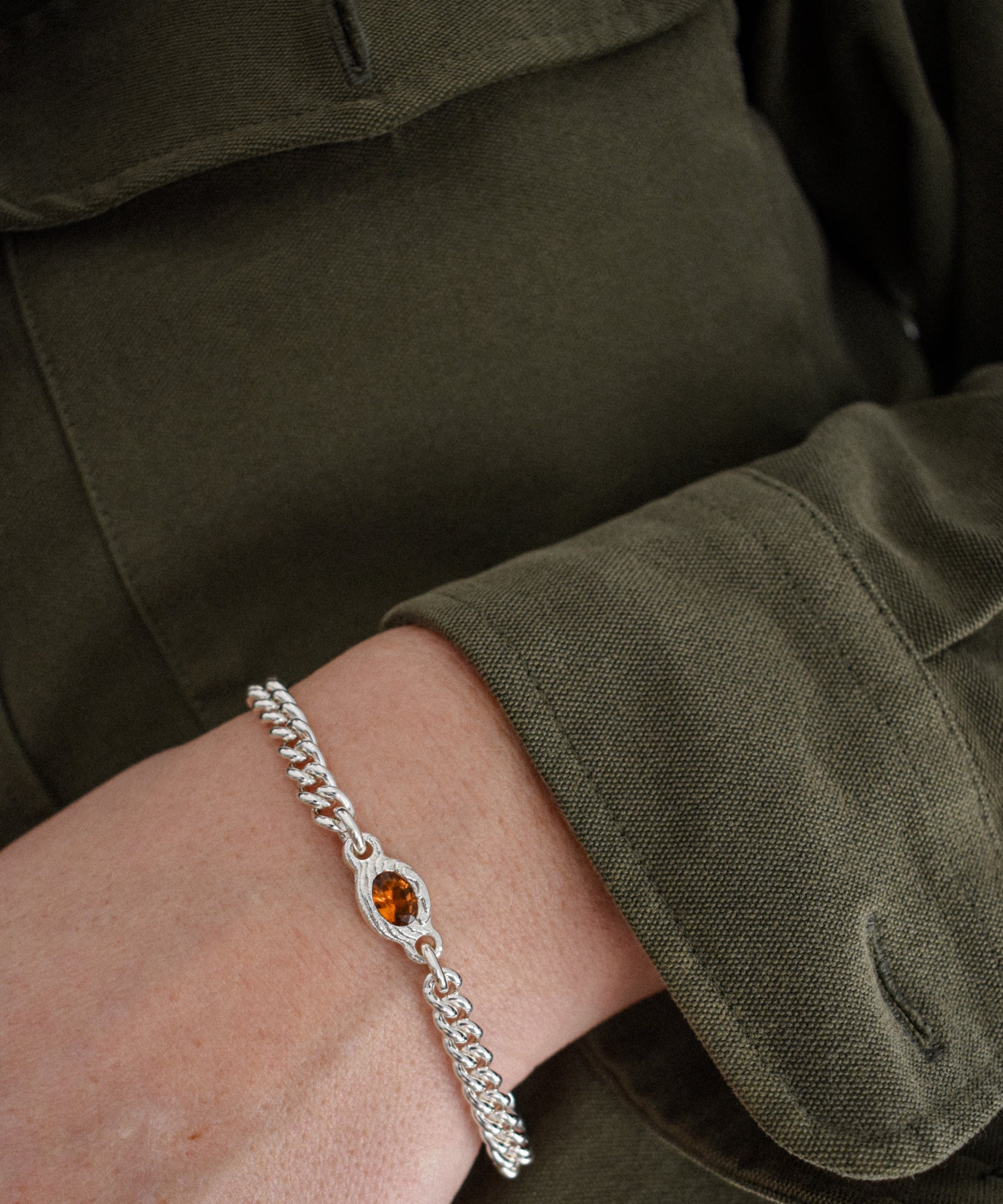 A close-up of a person's wrist wearing a silver chain bracelet with a central oval amber gemstone, set against an olive green military-style jacket.