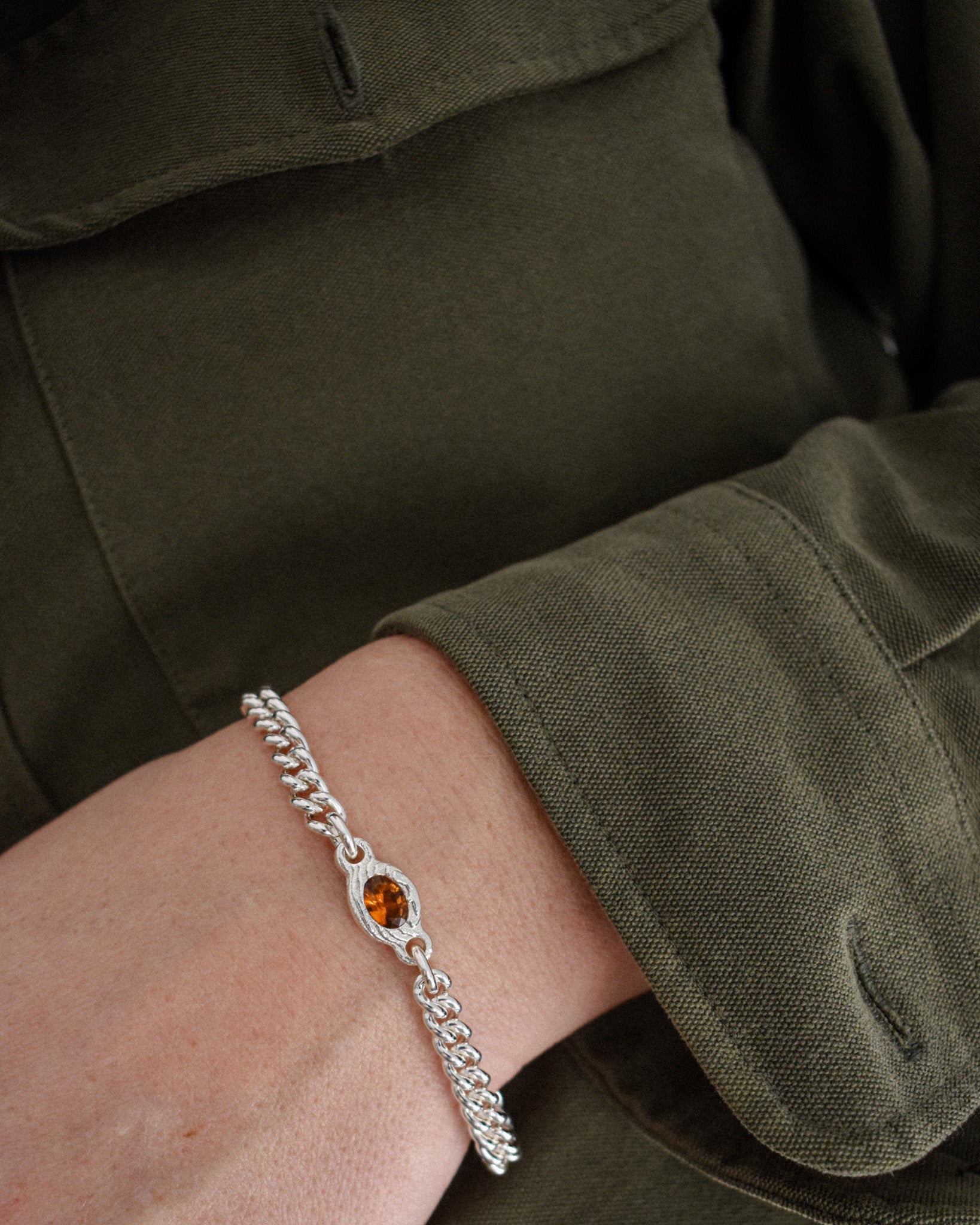 A close-up of a person's wrist wearing a silver chain bracelet with a central oval amber gemstone, set against an olive green military-style jacket.