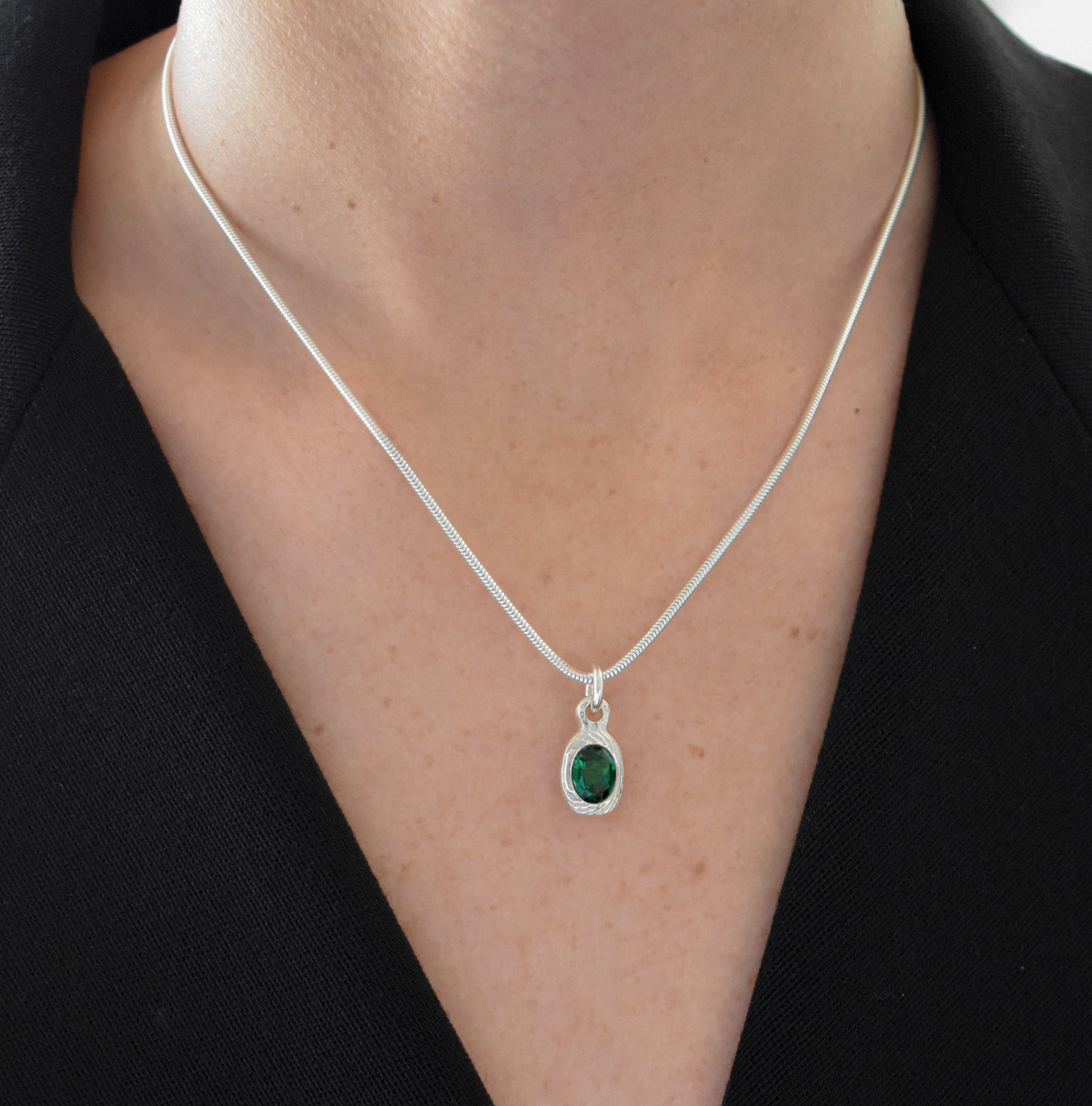 A close-up of a person's neck and chest wearing a delicate silver chain necklace with an oval green gemstone pendant set in a silver bezel.