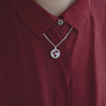 A close-up of a person wearing a silver chain necklace with a circular pendant featuring a red and white stylized 'S' logo, layered over a dark red collared shirt.