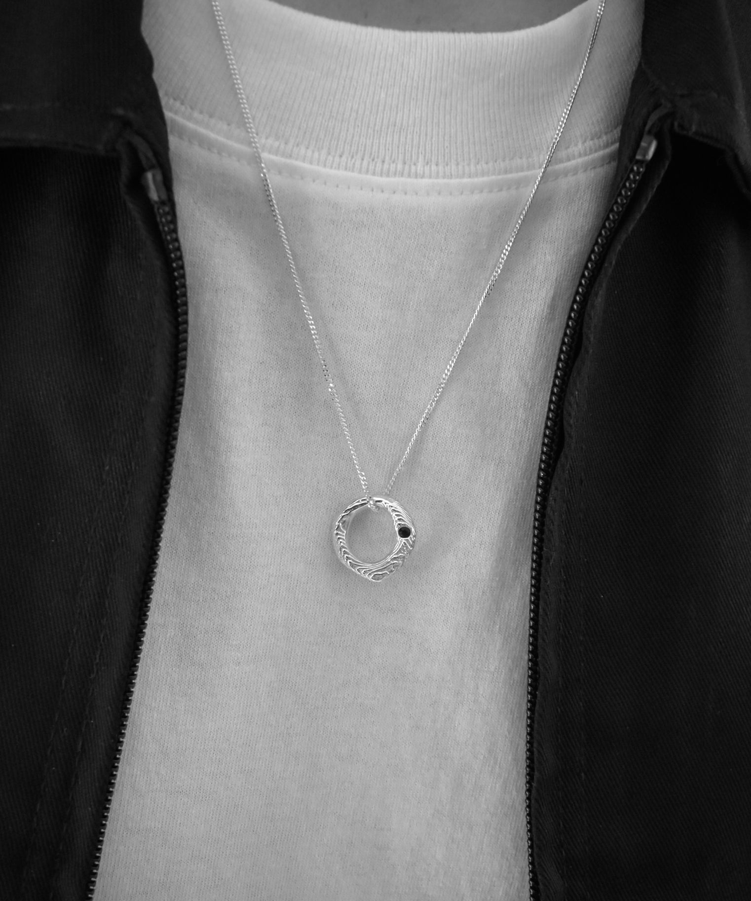A silver necklace with a textured circular pendant worn by a person in a black zip-up jacket over a white t-shirt.