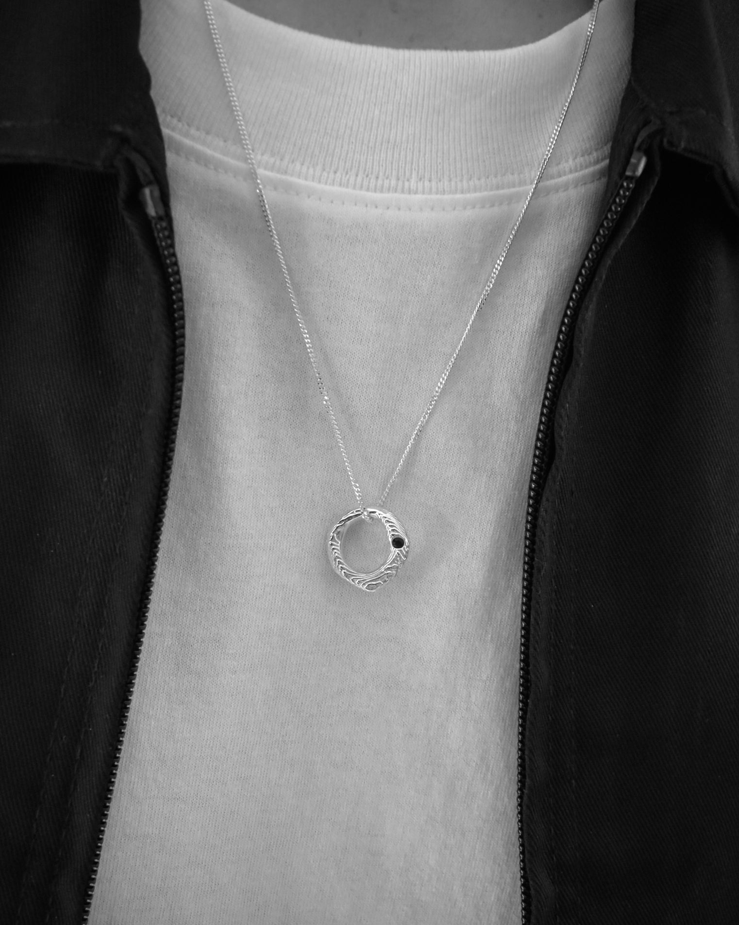 A silver necklace with a textured circular pendant worn by a person in a black zip-up jacket over a white t-shirt.