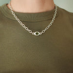 A close-up of a person wearing a silver chain necklace with a small oval green gemstone pendant, paired with an olive green crewneck shirt.