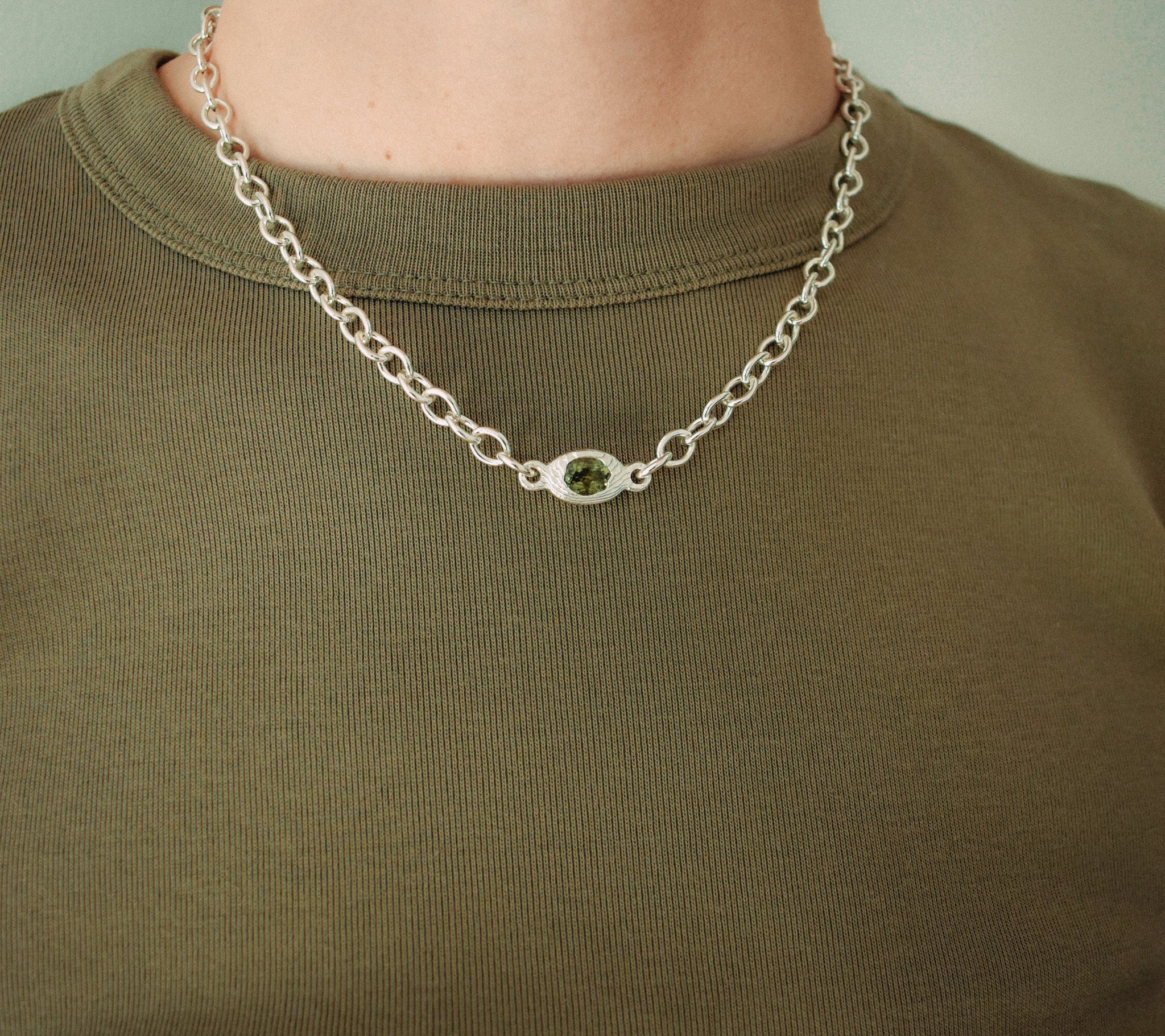 A close-up of a person wearing a silver chain necklace with a small oval green gemstone pendant, paired with an olive green crewneck shirt.