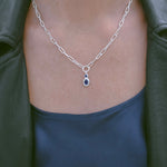 A close-up of a person wearing a silver paperclip chain necklace with a teardrop-shaped dark blue gemstone pendant, styled with a dark green leather jacket and a dark blue top.