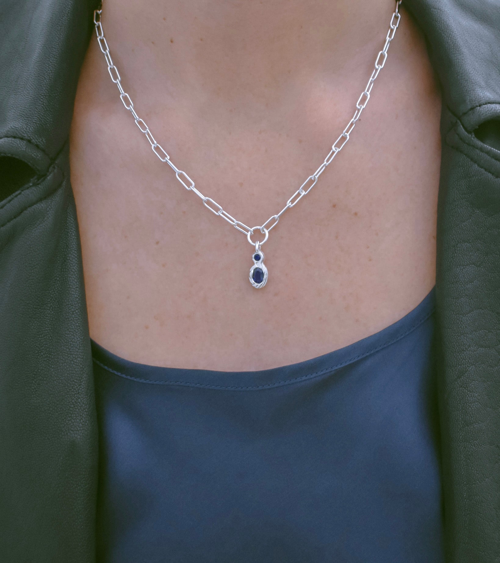 A close-up of a person wearing a silver paperclip chain necklace with a teardrop-shaped dark blue gemstone pendant, styled with a dark green leather jacket and a dark blue top.