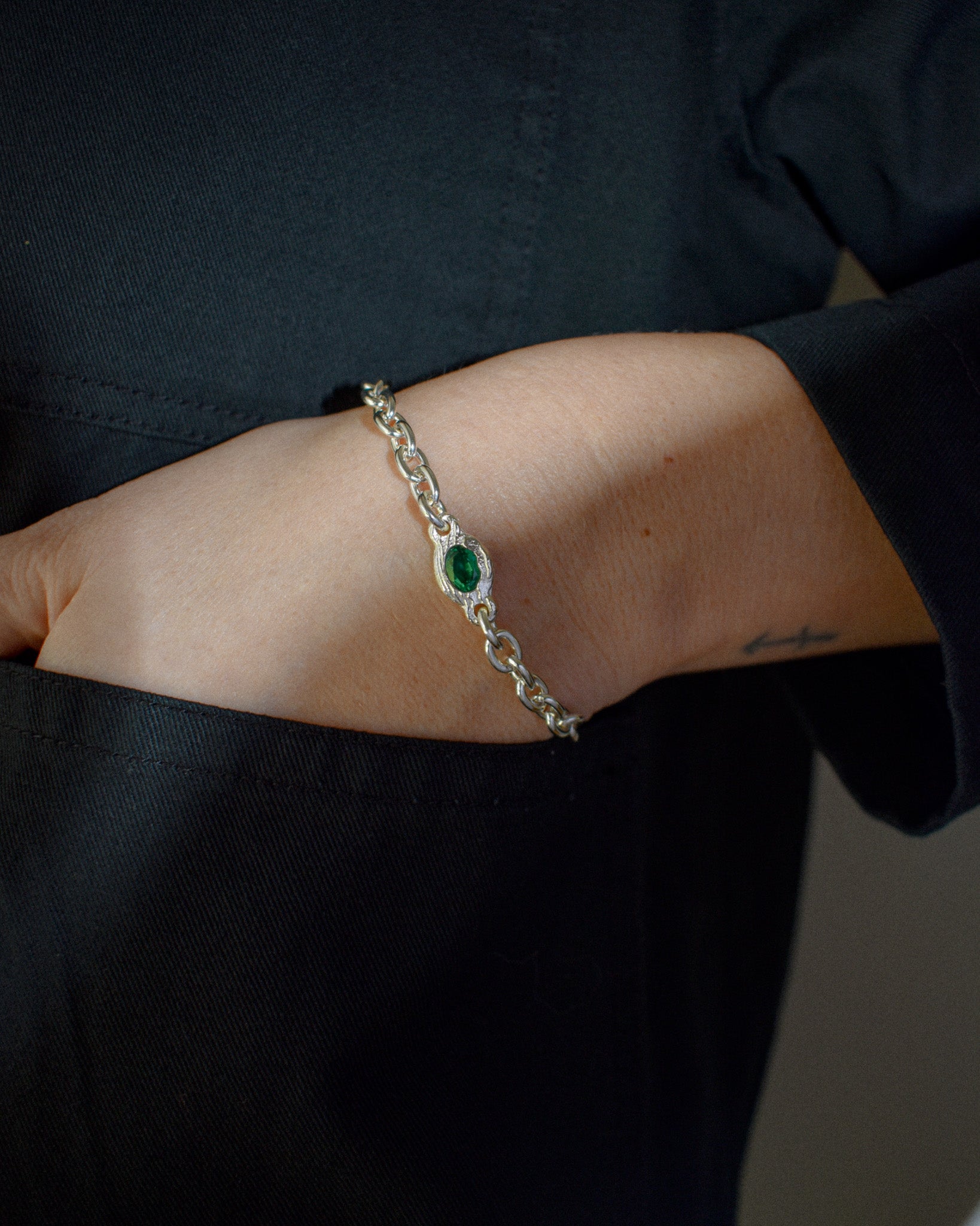 A close-up of a person's wrist wearing a silver chain bracelet with a central green oval stone, set against a dark navy shirt.