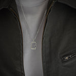 A silver rectangular pendant necklace with a small black dot detail, worn on a chain over a light gray t-shirt and dark gray zip-up jacket.