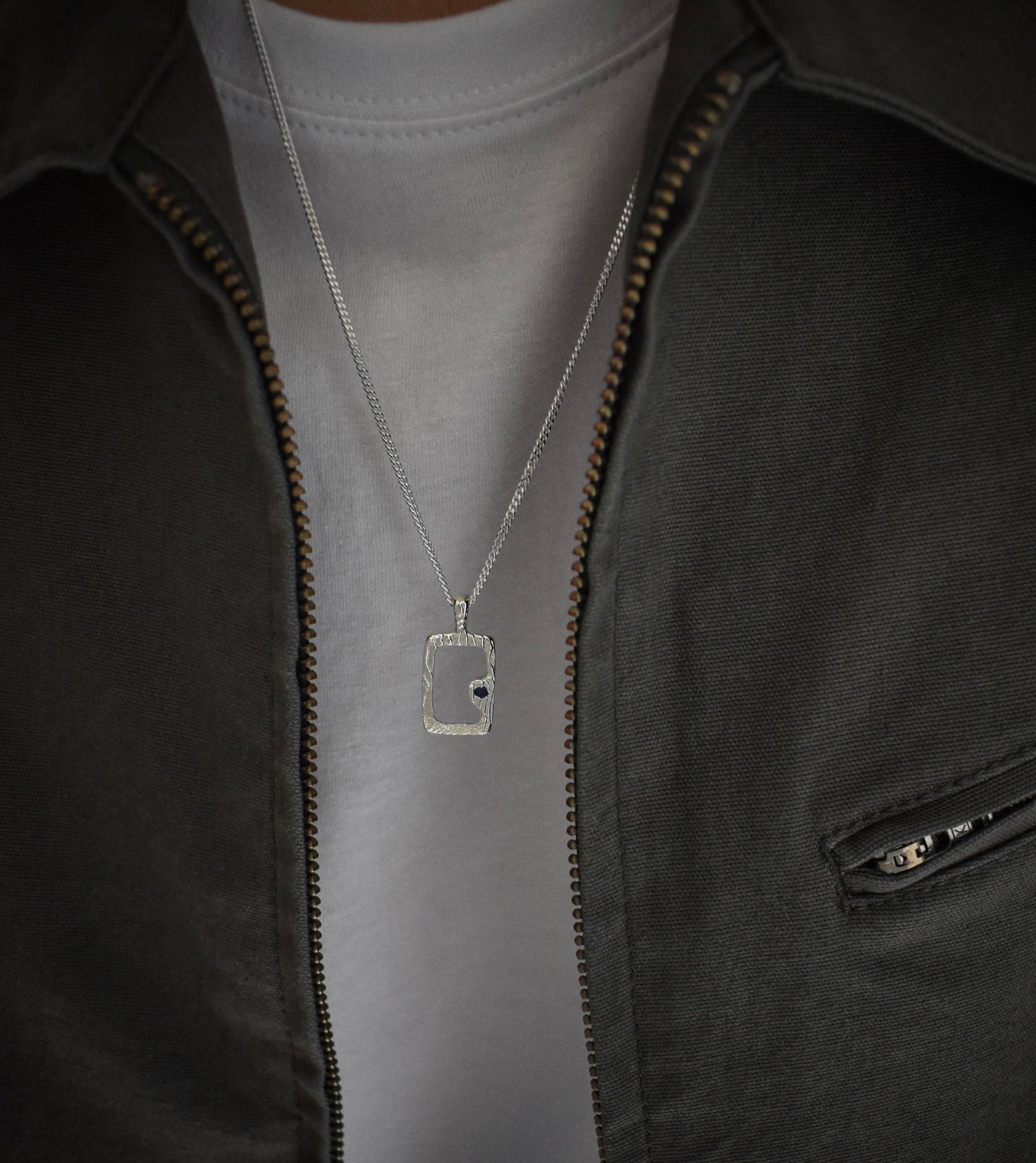 A silver rectangular pendant necklace with a small black dot detail, worn on a chain over a light gray t-shirt and dark gray zip-up jacket.