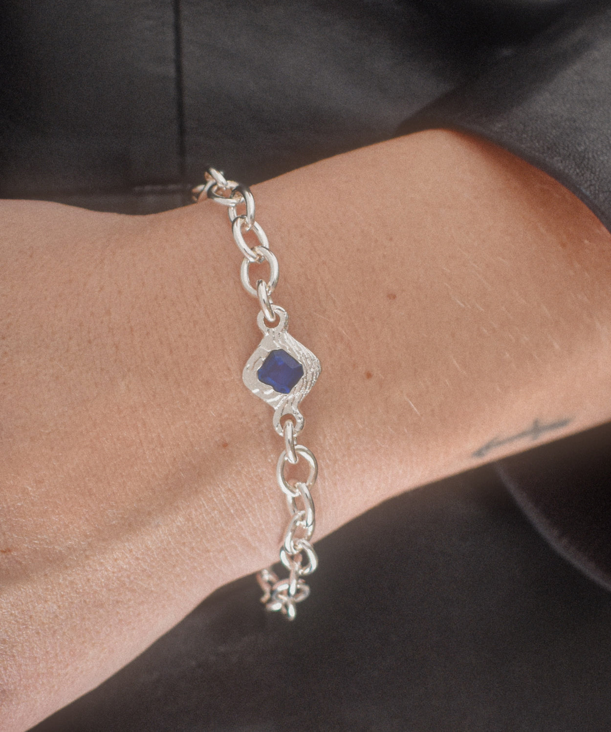 A sterling silver chain bracelet with a central diamond-shaped bezel holding a deep blue gemstone, shown worn on a person's wrist.