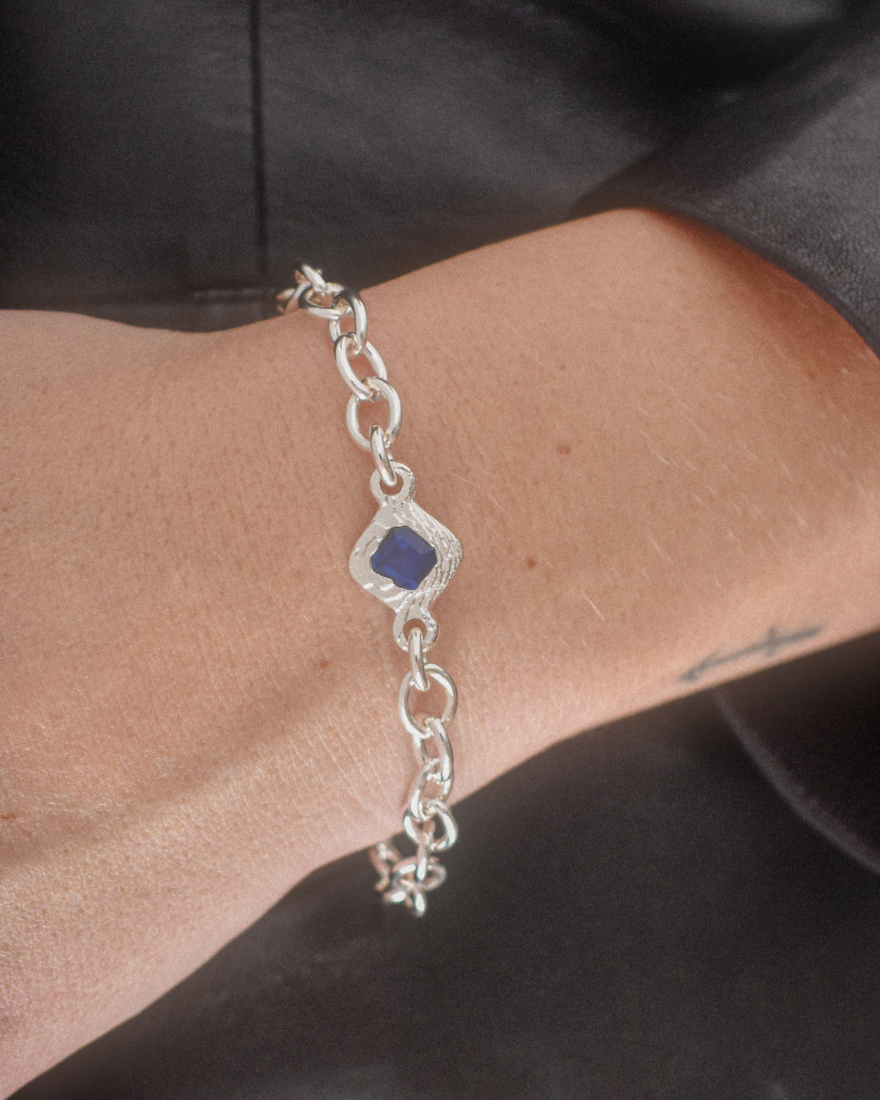 A sterling silver chain bracelet with a central diamond-shaped bezel holding a deep blue gemstone, shown worn on a person's wrist.