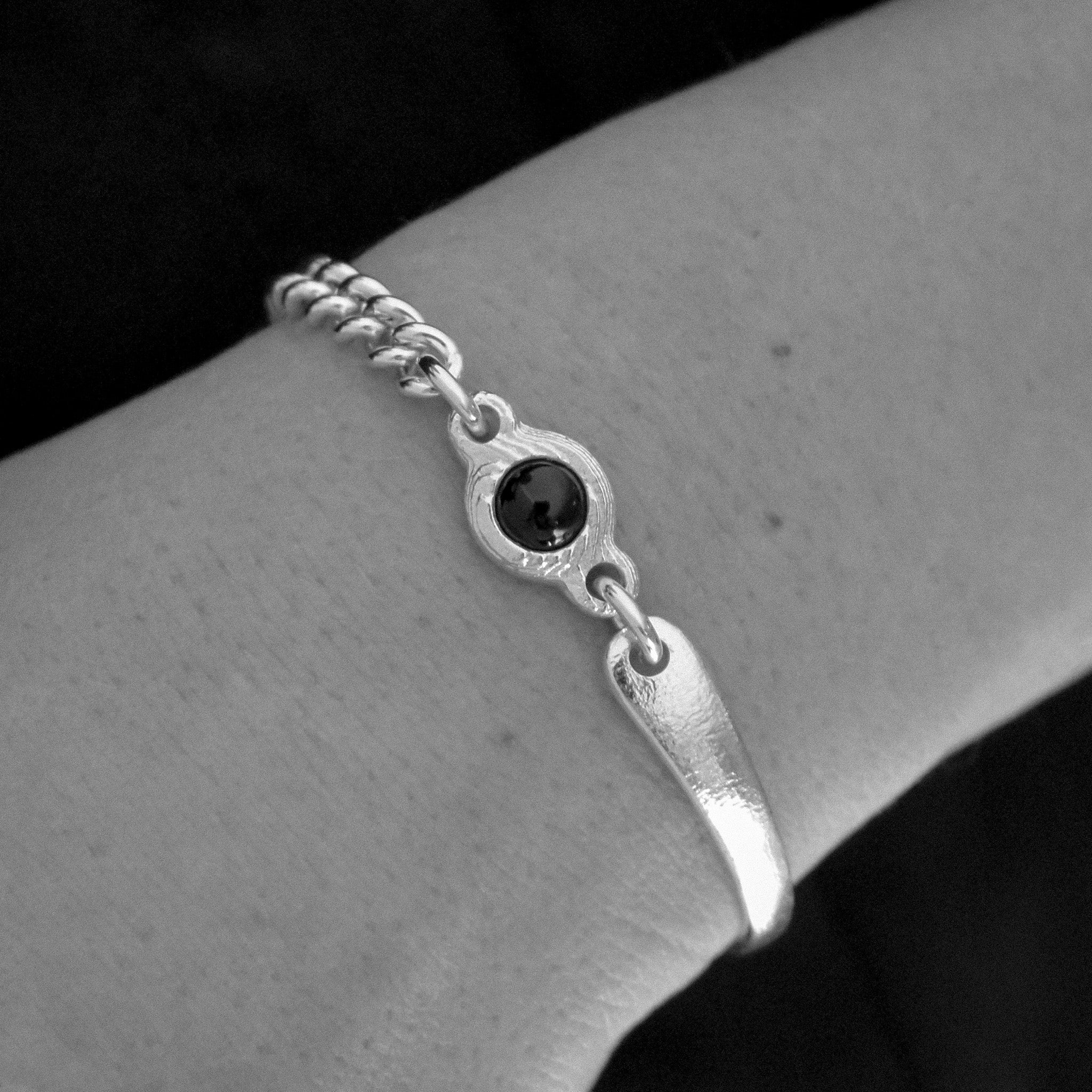 A silver-toned bangle bracelet with a textured chain, a central oval bezel containing a dark round stone, and a flat bar clasp, shown worn on a person's wrist.