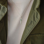 A silver dagger pendant necklace with a small green gemstone accent on the hilt, worn on a model's neck over a white t-shirt and olive green jacket.