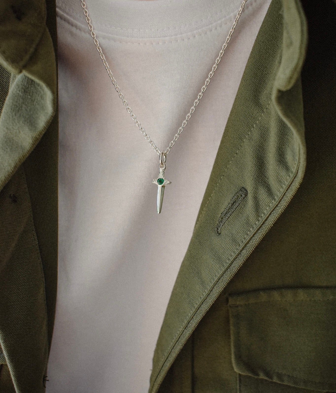 A silver dagger pendant necklace with a small green gemstone accent on the hilt, worn on a model's neck over a white t-shirt and olive green jacket.