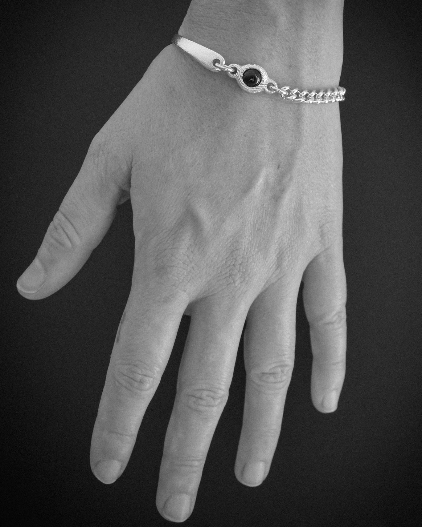 A silver chain bracelet with a central black oval stone, shown worn on a hand against a dark gray background.