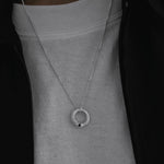 A silver-toned chain necklace with a textured circular pendant featuring a small black dot, worn by a person over a light gray crewneck sweatshirt and a black zip jacket.