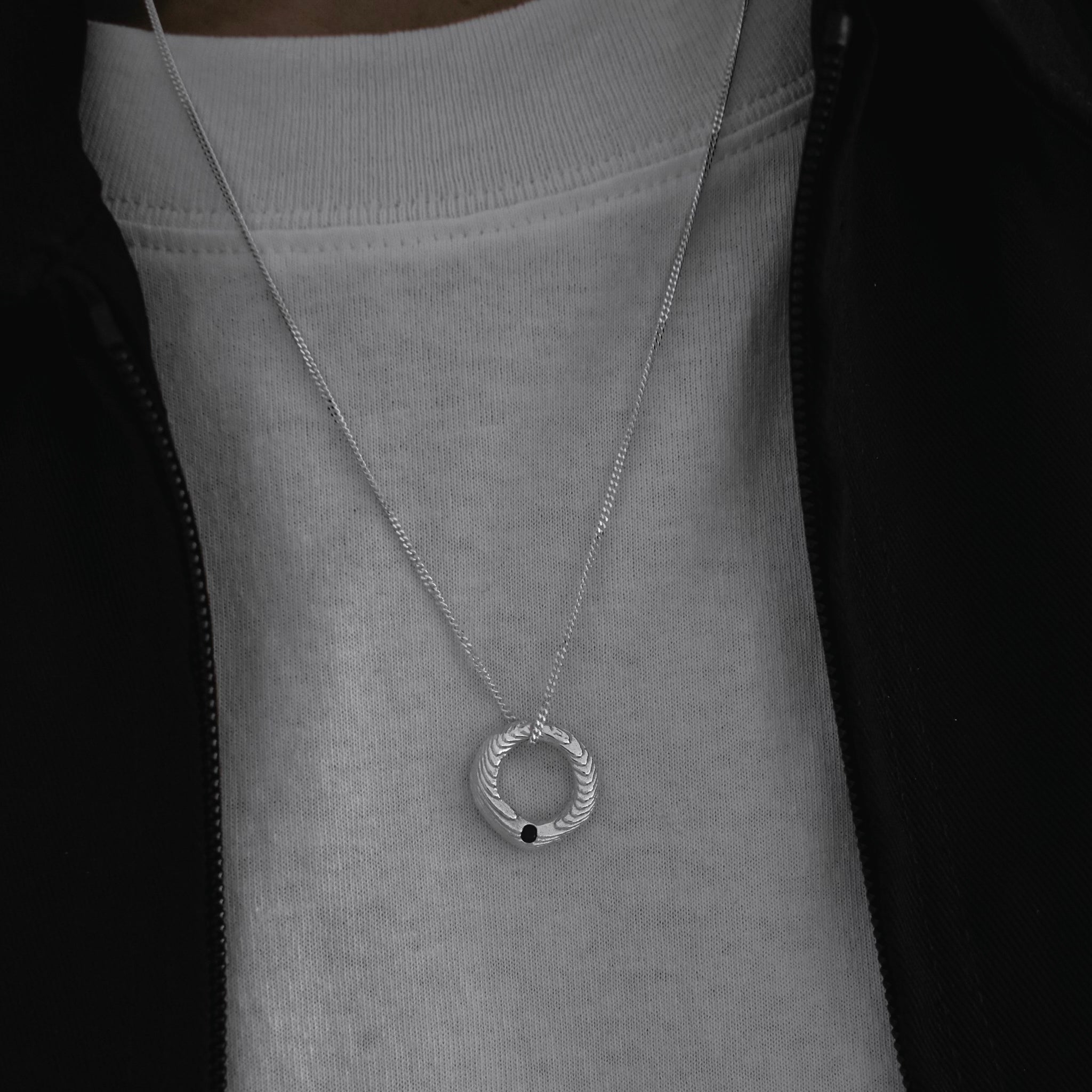 A silver-toned chain necklace with a textured circular pendant featuring a small black dot, worn by a person over a light gray crewneck sweatshirt and a black zip jacket.