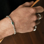 A close-up of a person's hand wearing a silver chain bracelet with a small green oval stone, alongside silver rings, set against a dark background.