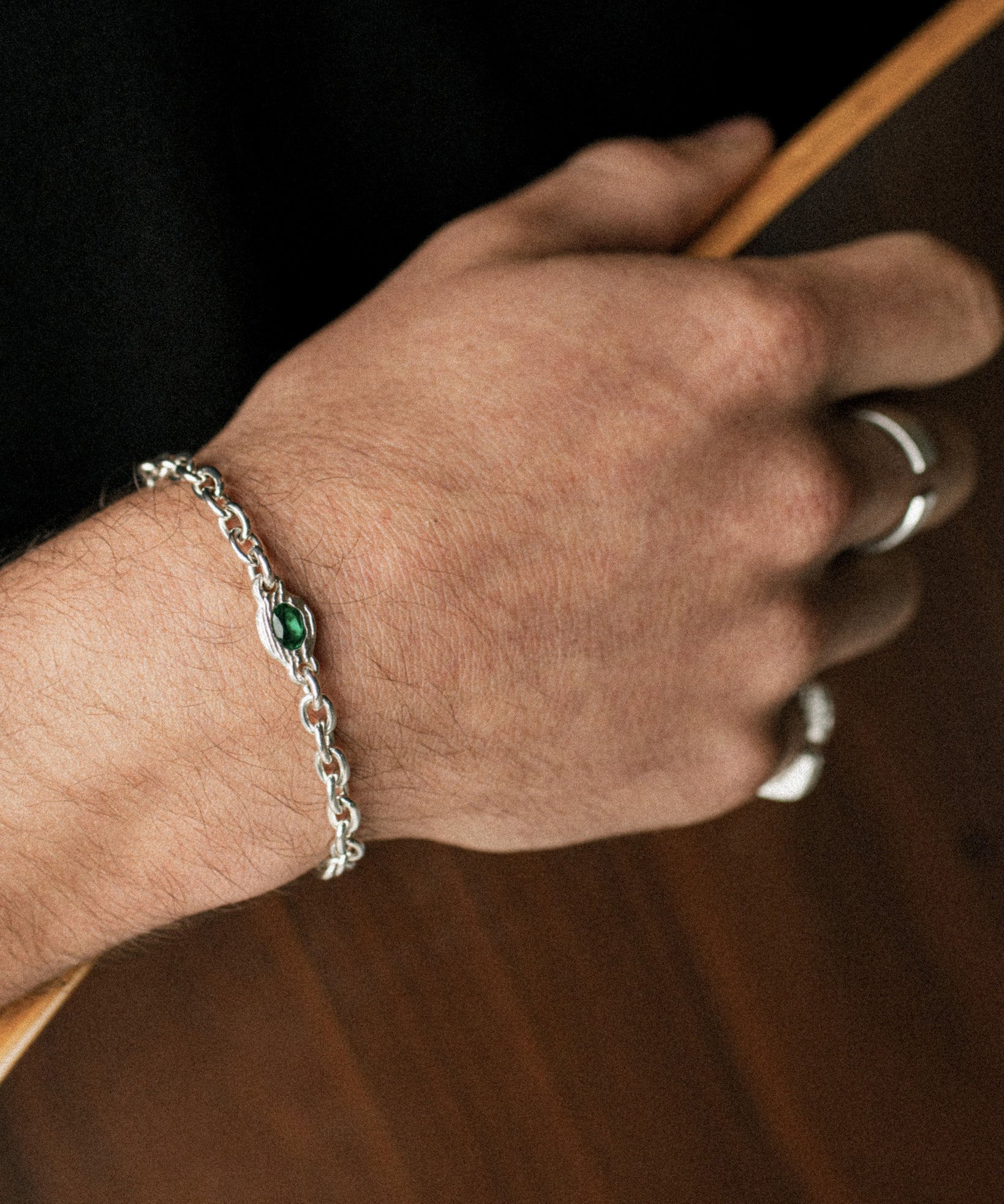 A close-up of a person's hand wearing a silver chain bracelet with a small green oval stone, alongside silver rings, set against a dark background.