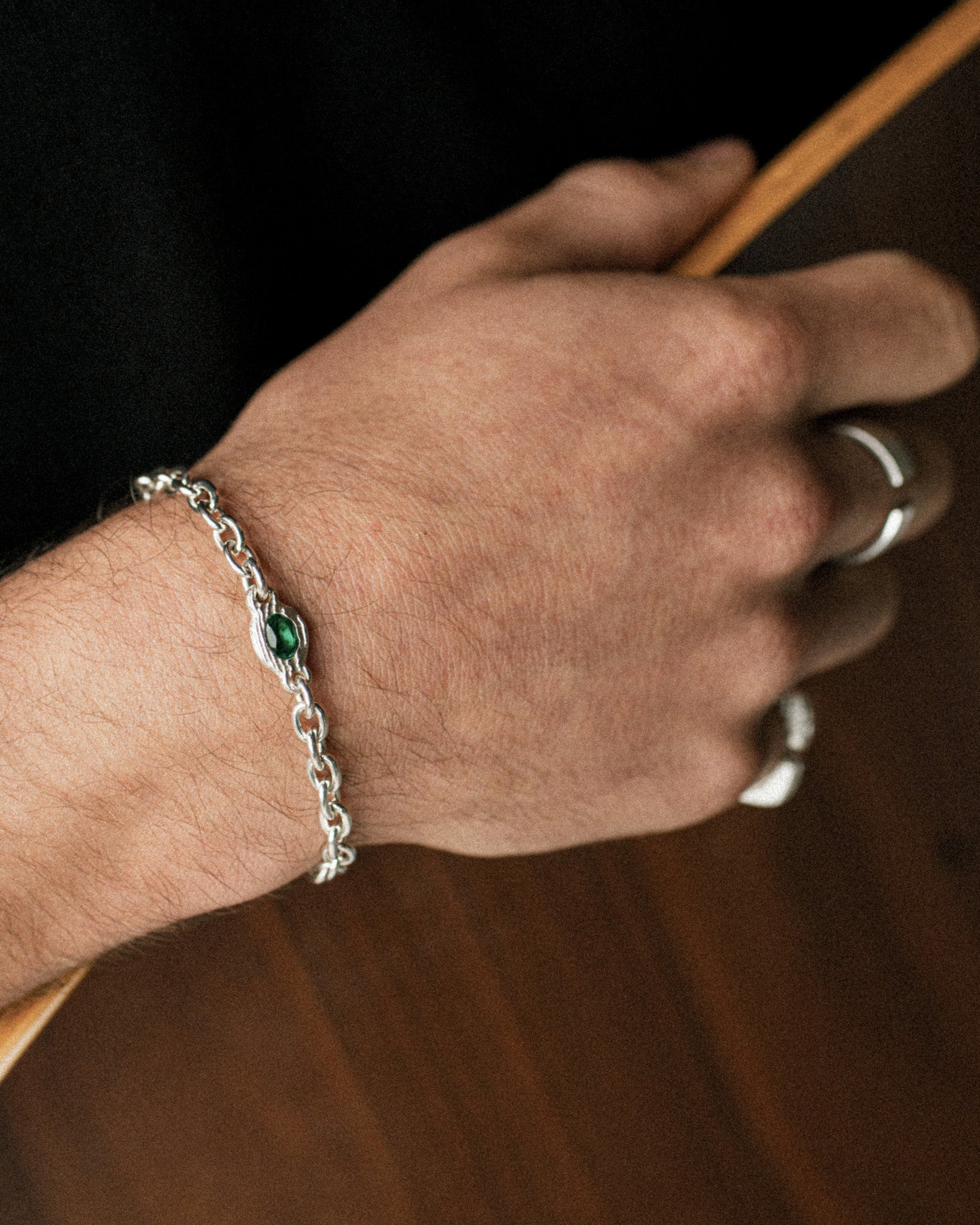 A close-up of a person's hand wearing a silver chain bracelet with a small green oval stone, alongside silver rings, set against a dark background.