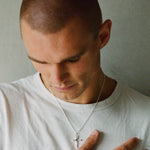 A close-up shot of a man with a shaved head wearing a white t-shirt and a silver cross pendant necklace, looking down with his hand resting on his chest against a plain gray wall.