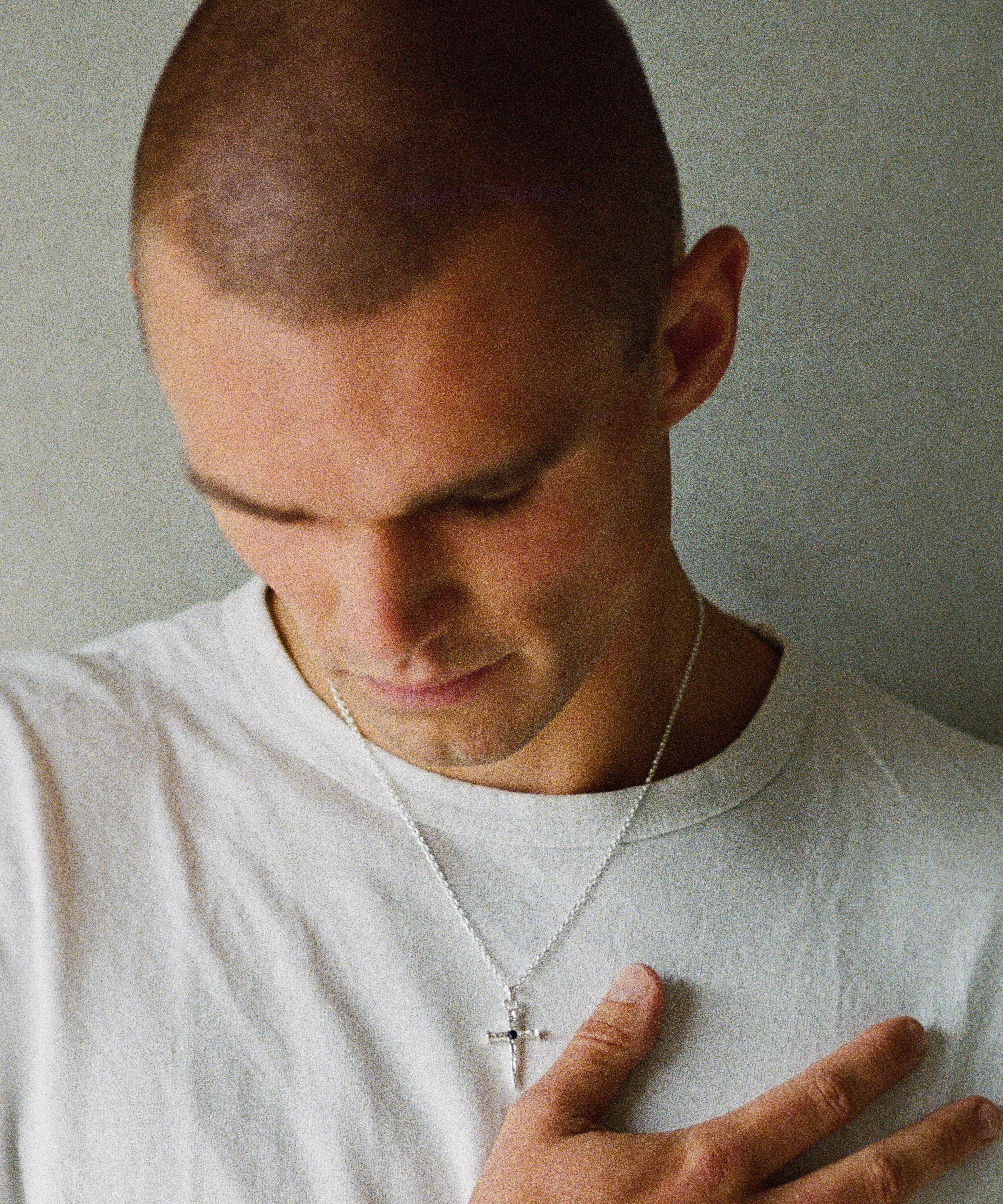A close-up shot of a man with a shaved head wearing a white t-shirt and a silver cross pendant necklace, looking down with his hand resting on his chest against a plain gray wall.
