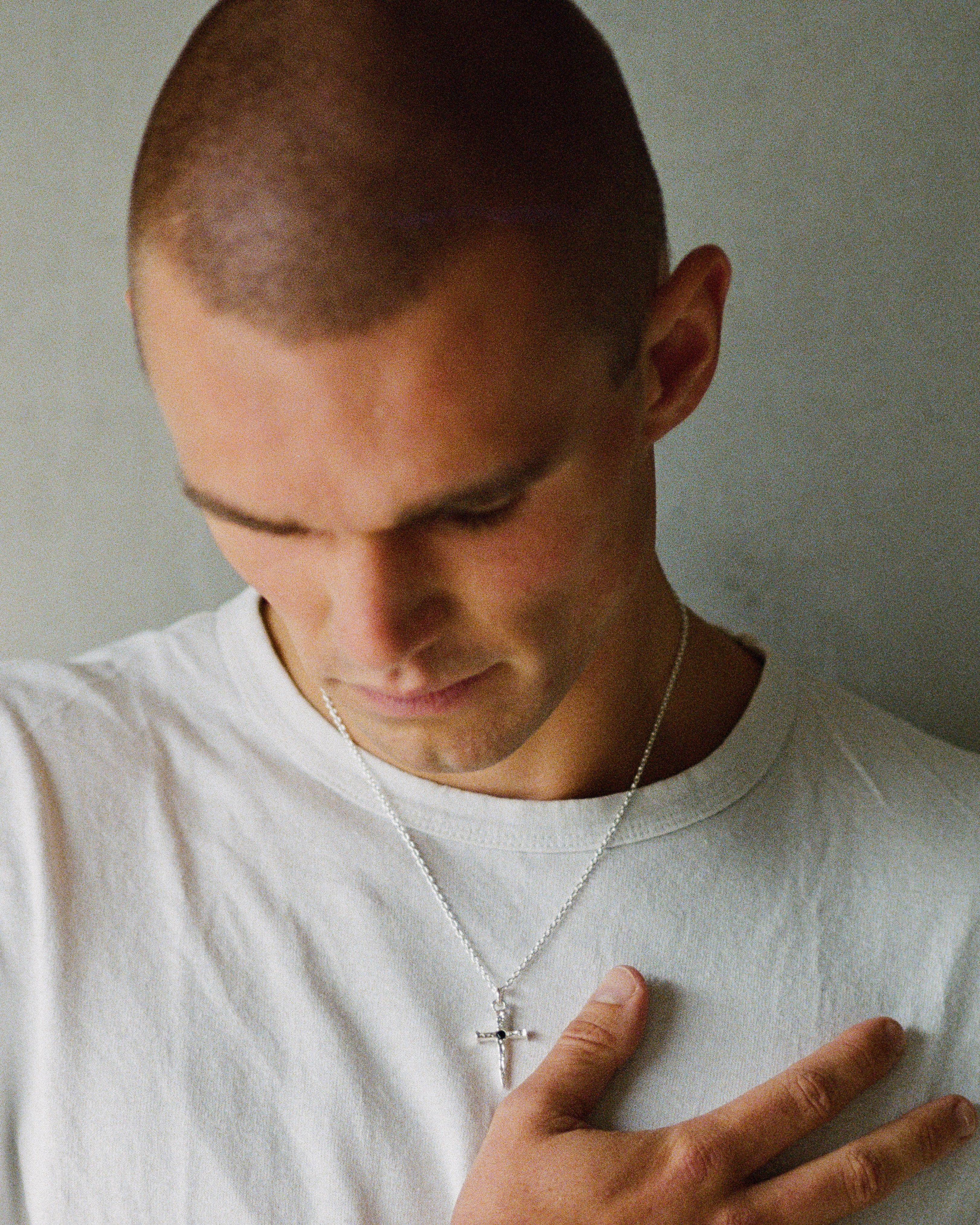 A close-up shot of a man with a shaved head wearing a white t-shirt and a silver cross pendant necklace, looking down with his hand resting on his chest against a plain gray wall.