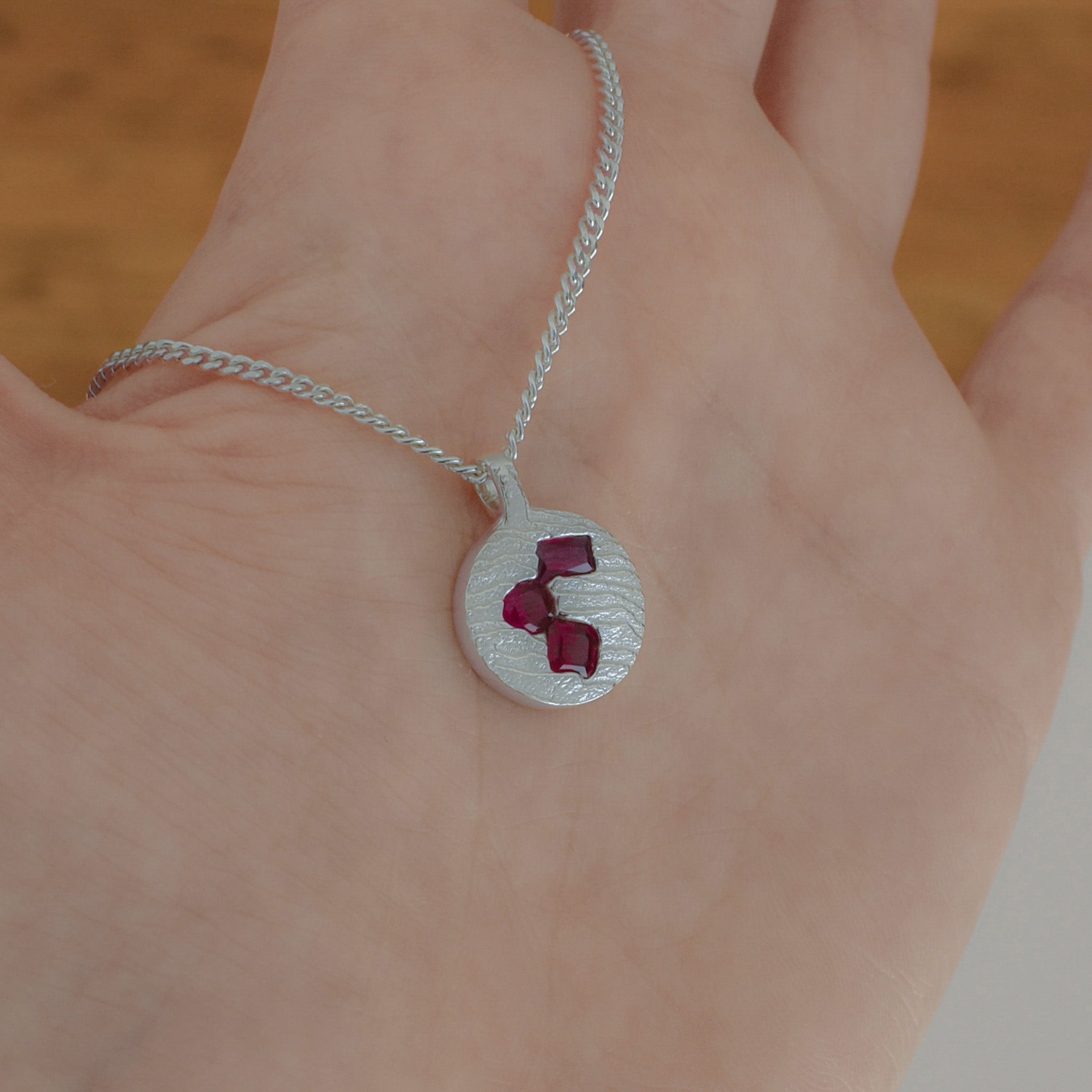 A close-up of a hand wearing a delicate silver chain necklace with a textured silver oval pendant featuring three small red gemstones.