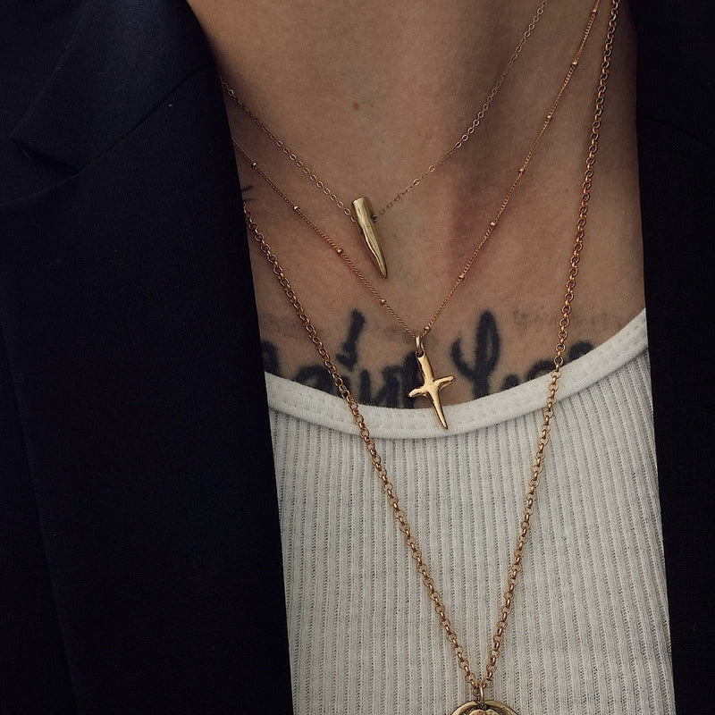 Close-up of layered gold necklaces with a cross pendant on a person wearing a black jacket and white shirt.
