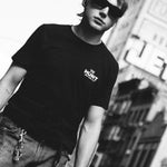 A man wearing a black t-shirt with a white 'THE HUNT' logo on the chest, styled with distressed jeans and sunglasses, set against a blurred urban background.