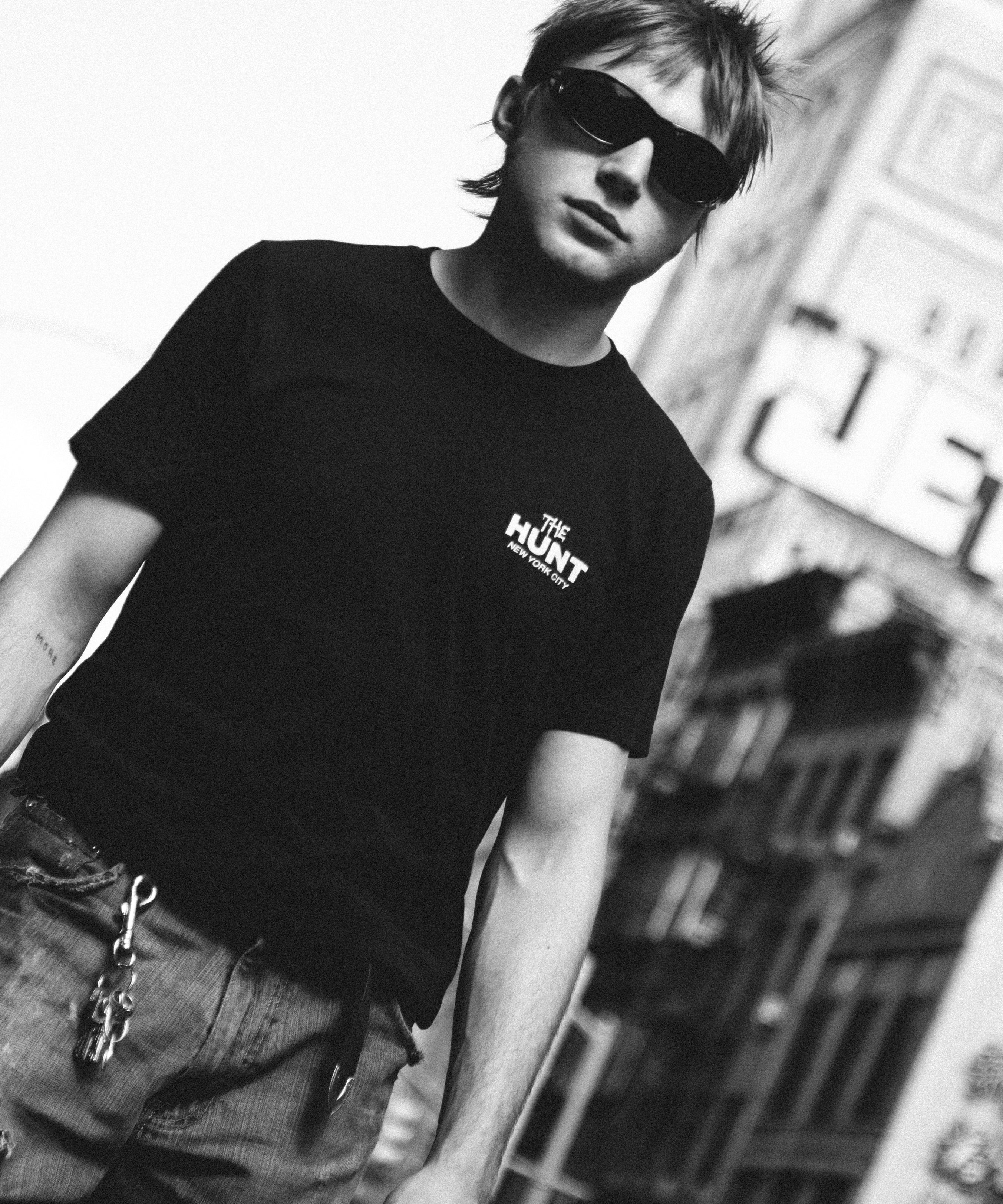 A man wearing a black t-shirt with a white 'THE HUNT' logo on the chest, styled with distressed jeans and sunglasses, set against a blurred urban background.