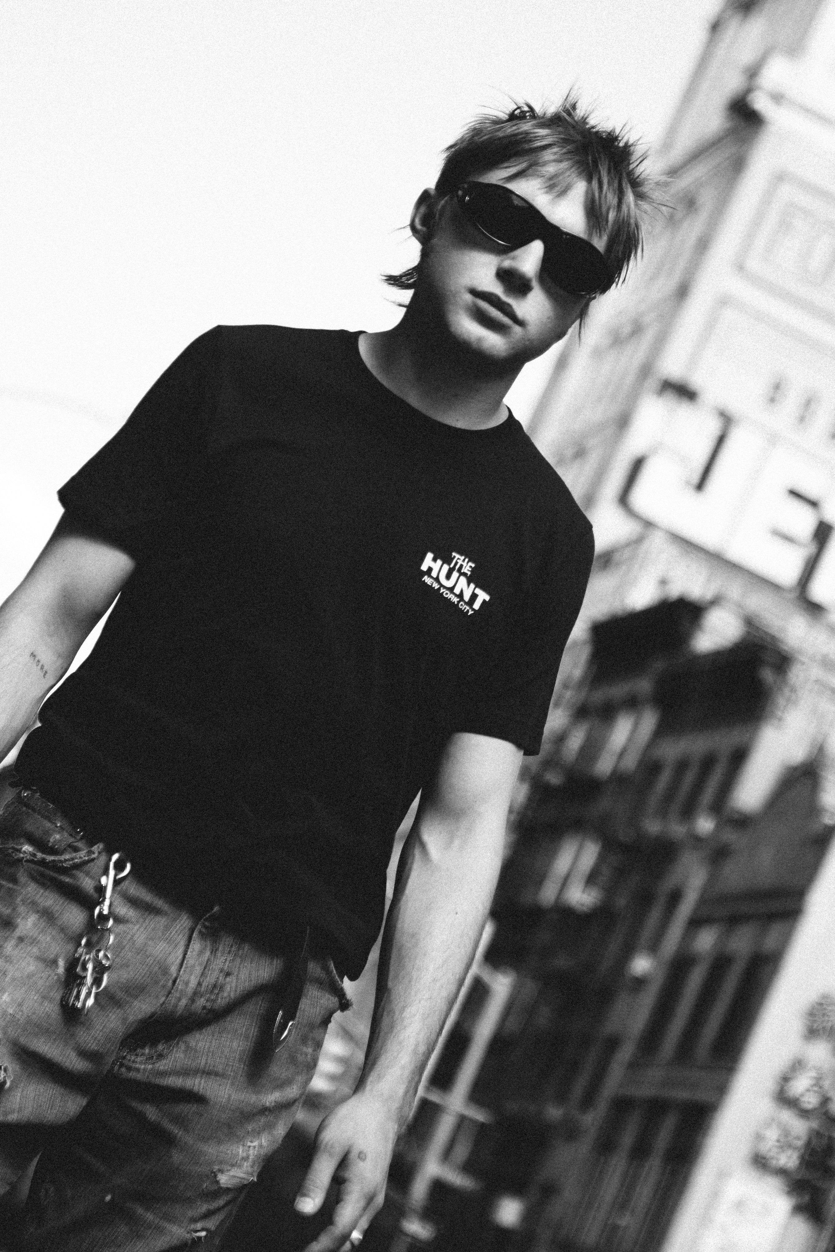 A man wearing a black t-shirt with a white 'THE HUNT' logo on the chest, styled with distressed jeans and sunglasses, set against a blurred urban background.
