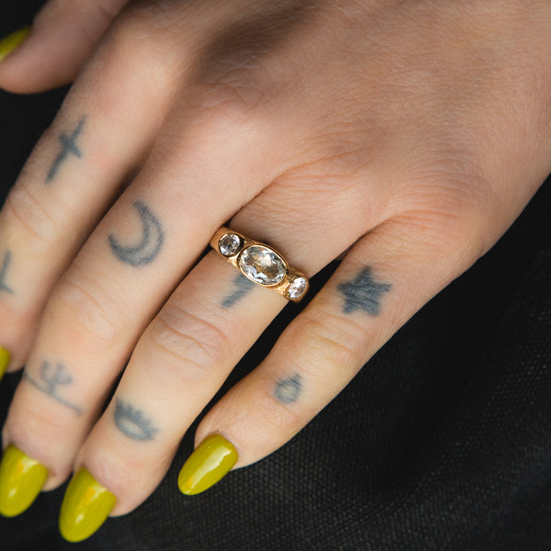 Hand with a gold ring featuring a large gemstone, green nail polish, and tattoos on a dark background.